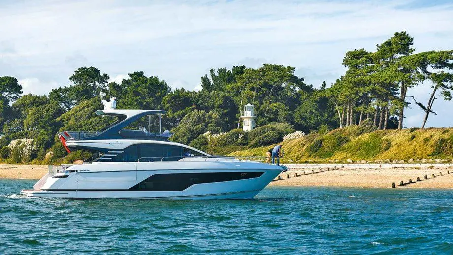 Luxury motor yacht cruising near a sandy shore, with a man on the beach and a lighthouse surrounded by trees in the background.